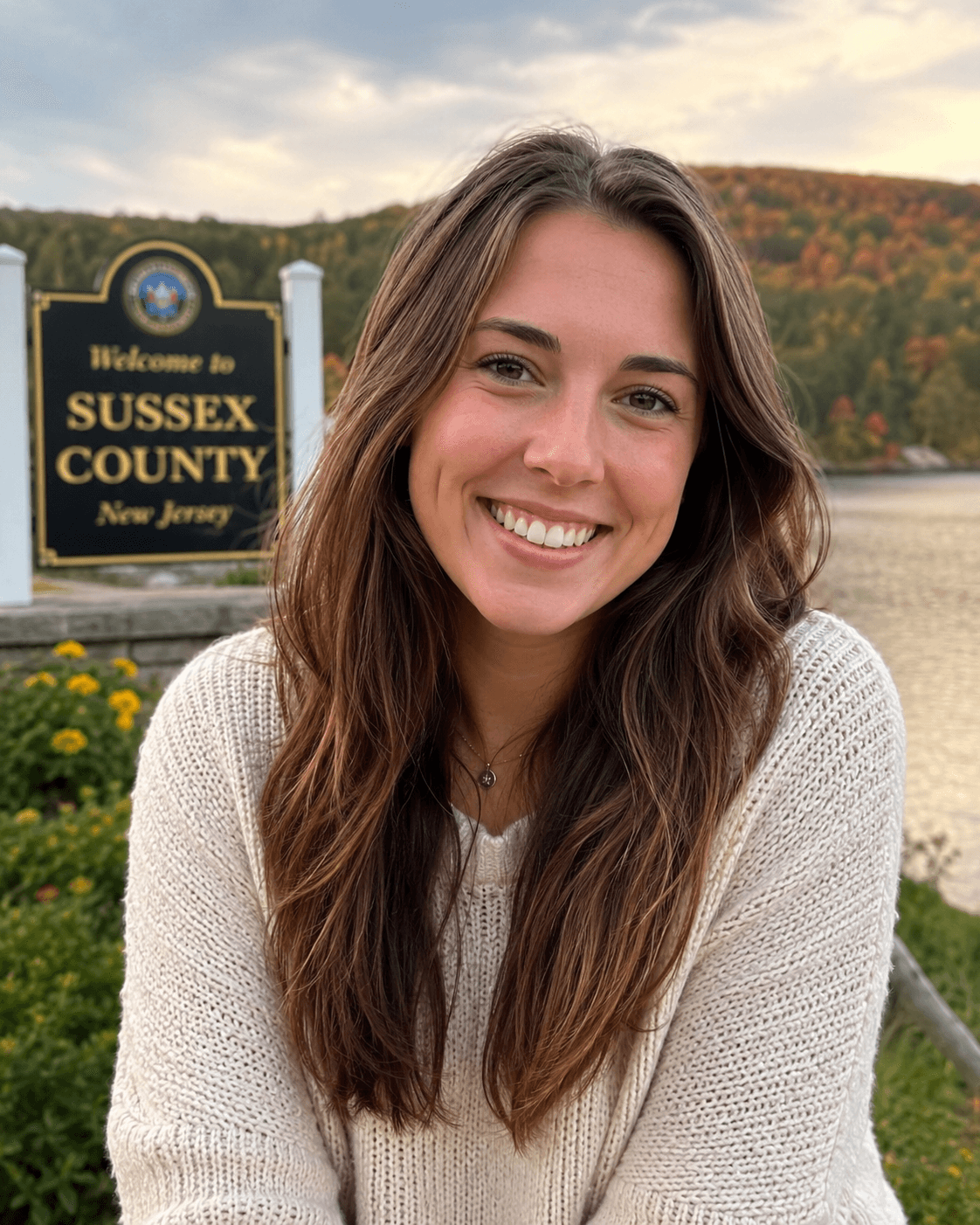 A Sussex County, New Jersey resident at the Welcome to Sussex County sign, with the autumn hills and lake behind