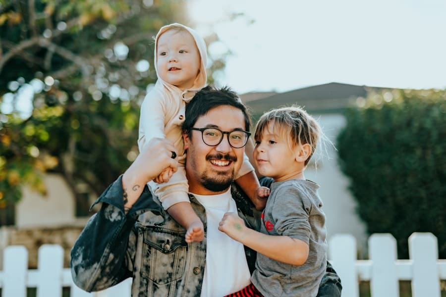 A smiling father outside his home carrying his two young children